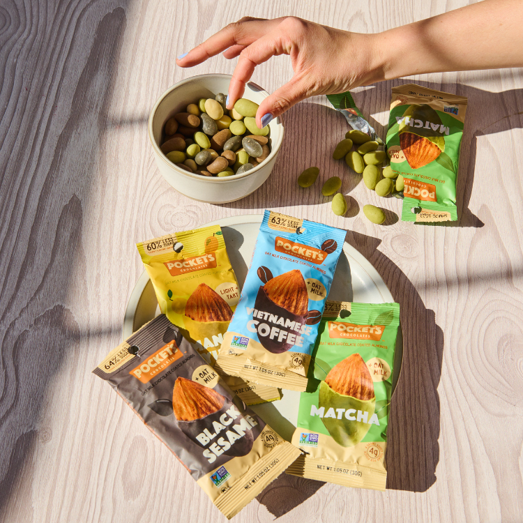 A hand reaches for a bowl of mixed almonds, with snack packs of Black Sesame, Matcha, and Vietnamese Coffee on a table