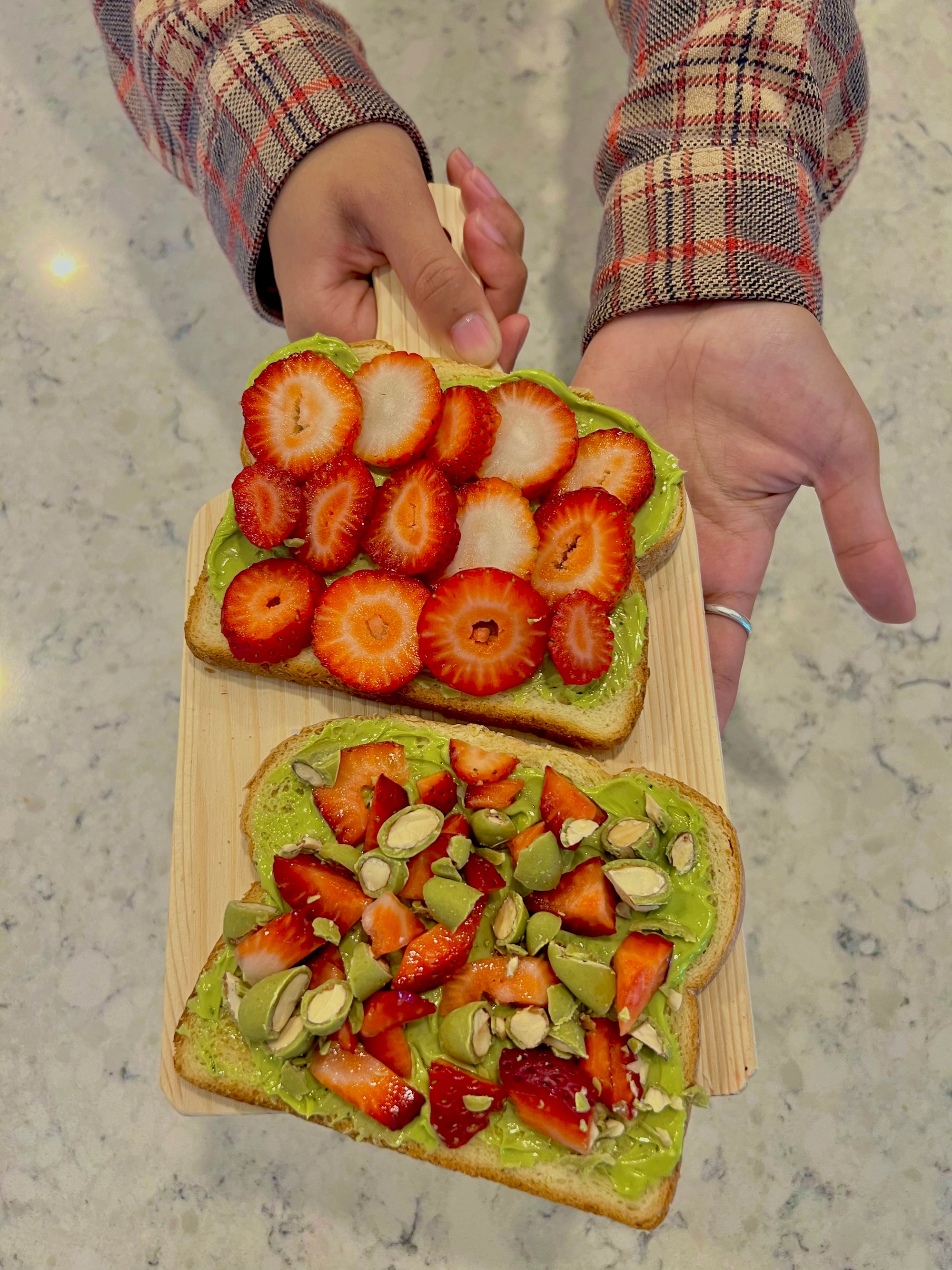 Hands presenting a wooden board with two slices of avocado toast topped with strawberries and seeds, over a marble surface
