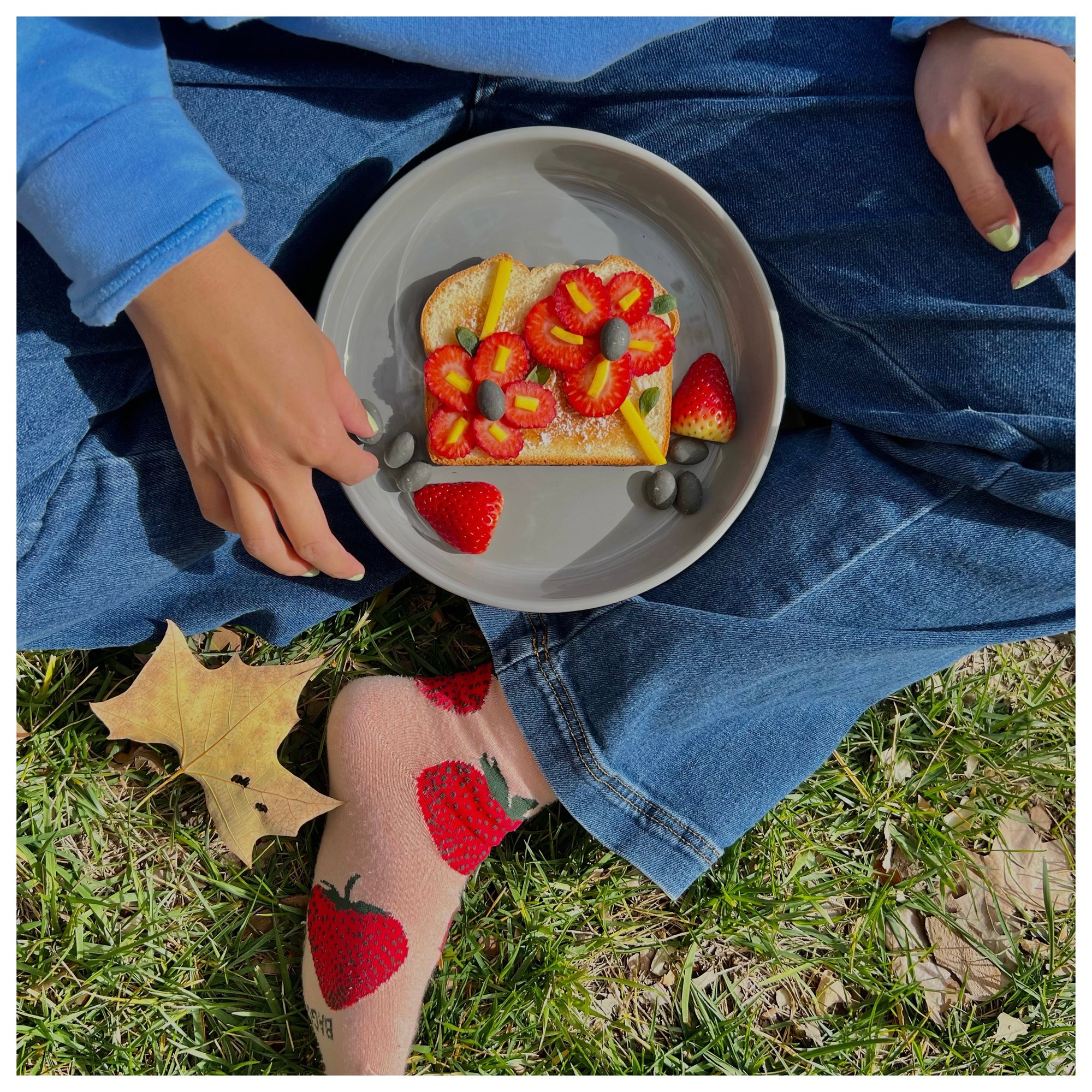 Person sitting on grass with a plate of fruit-topped toast, wearing jeans and strawberry socks